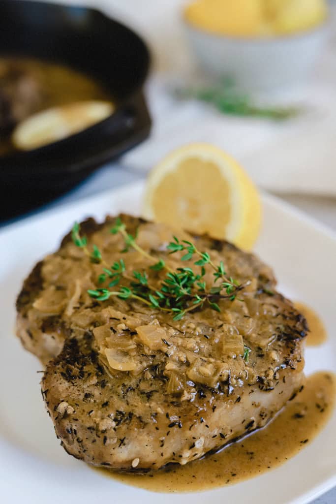 A pork chop topped with lemon garlic cream sauce on a white plate with a lemon half