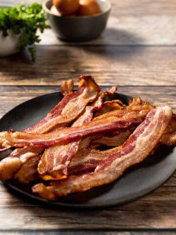 A plate of crispy oven bacon on a wood background next to eggs and parsley