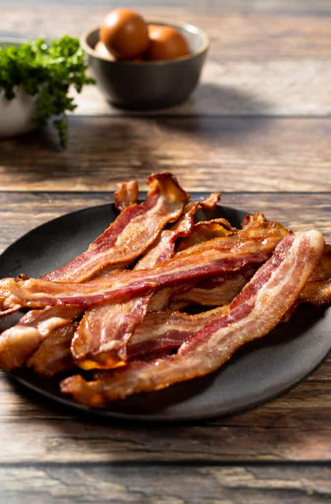 A plate of crispy oven bacon on a wood background next to eggs and parsley