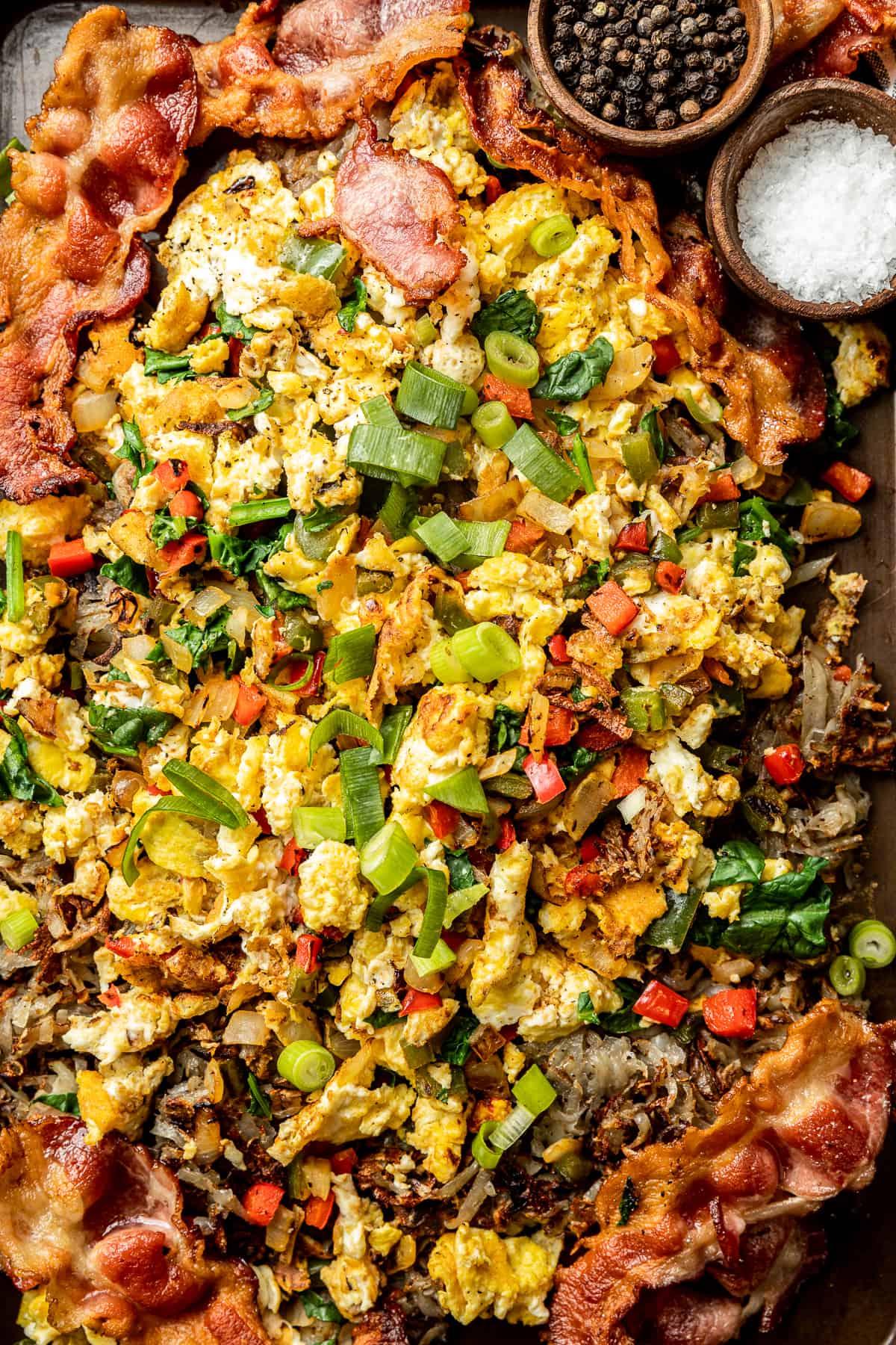 An overview shot of eggs, hashbrowns, and bacon on a baking sheet topped with green onions, salt, and pepper.