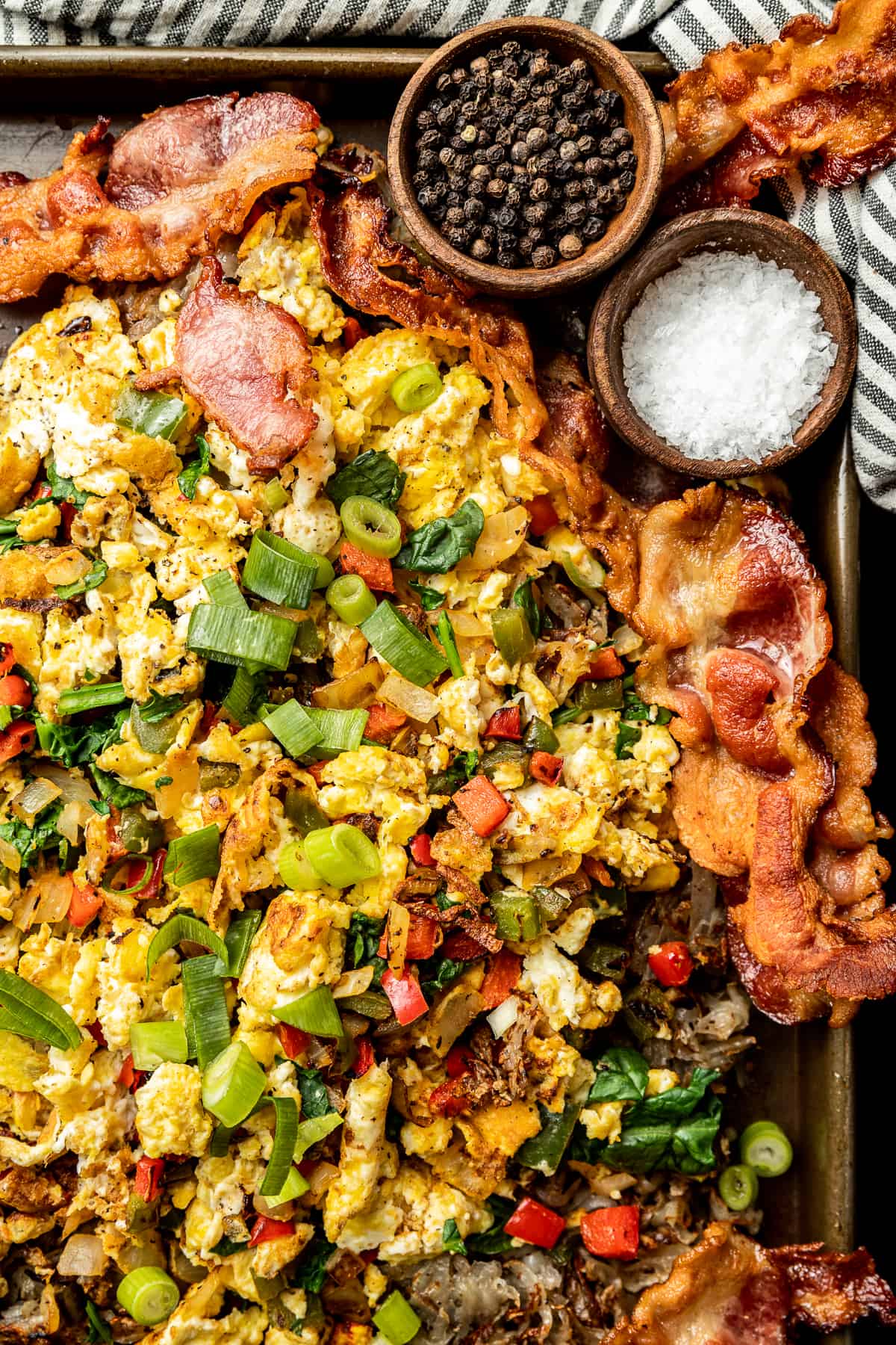 An overview shot of eggs, hashbrowns, and bacon on a baking sheet topped with green onions, salt, and pepper.