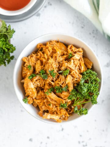 An overview shot of a bowl of buffalo chicken on a grey background next to greens and buffalo sauce