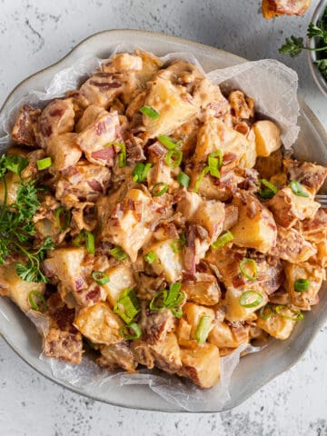 An overview shot of a bowl of buffalo ranch potato salad next to a bowl of parsley, bacon, and a towel