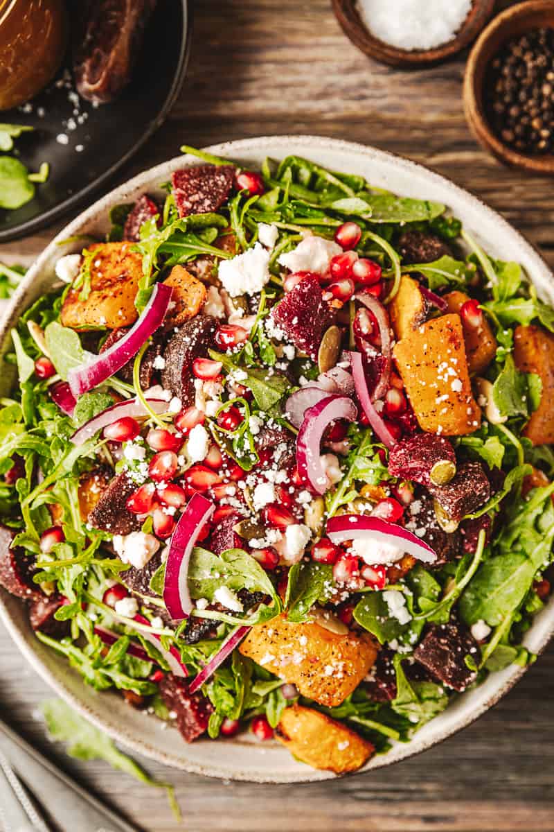 An overview shot of a bowl of a roasted butternut and beetroot salad on a wood background near utinsels and dressing