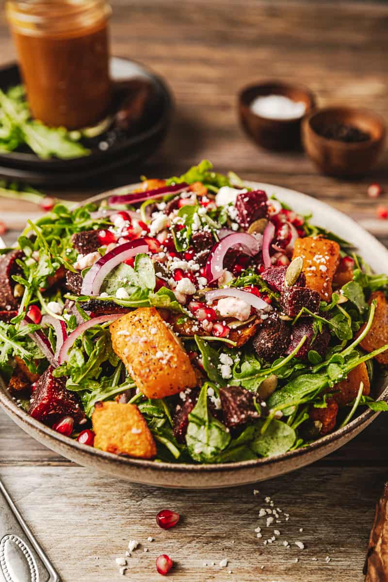 A prepared bowl of roasted butternut and beetroot salad on a wood background near feta cheese and pomegranate seeds