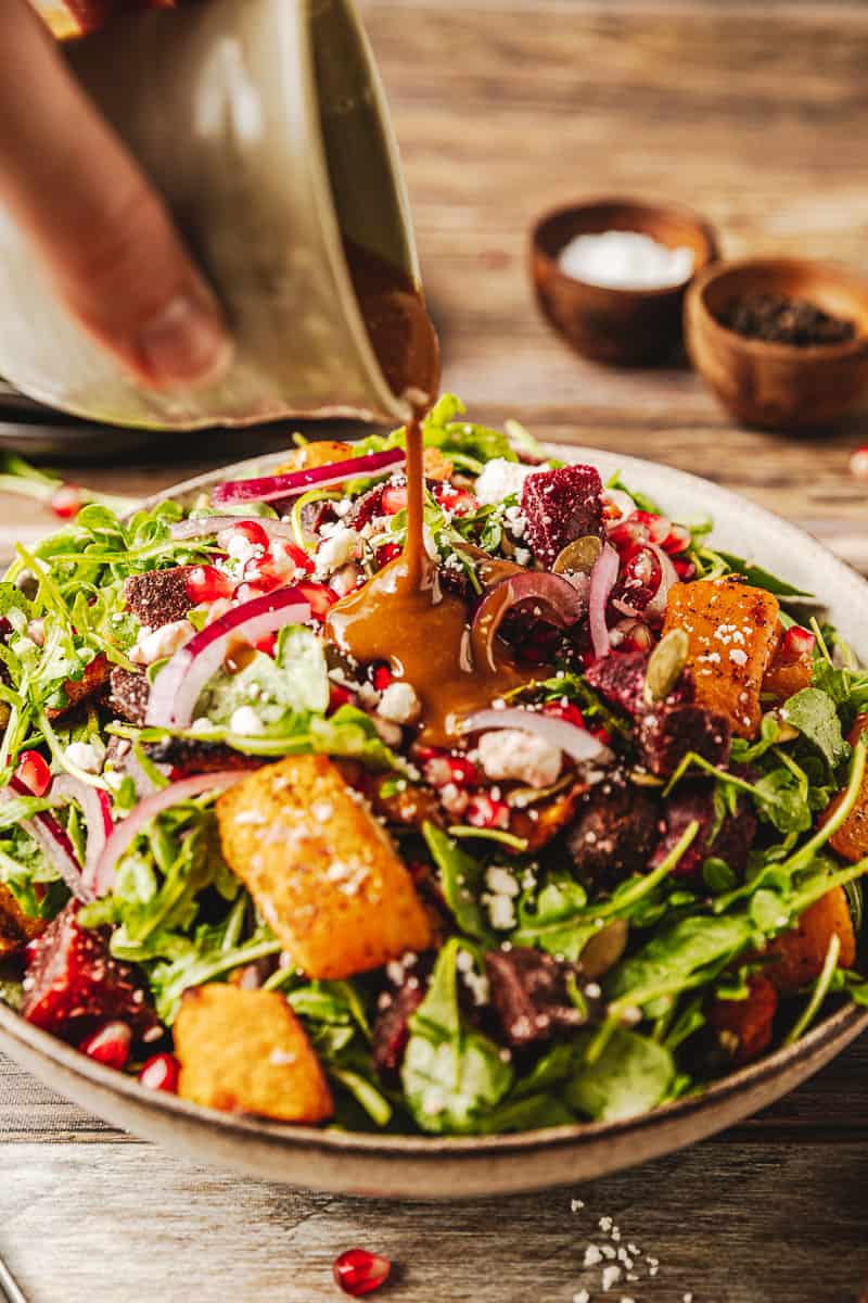 A ceramic jar of date and dijon dressing being poured onto the a bowl of roasted butternut and beet salad.