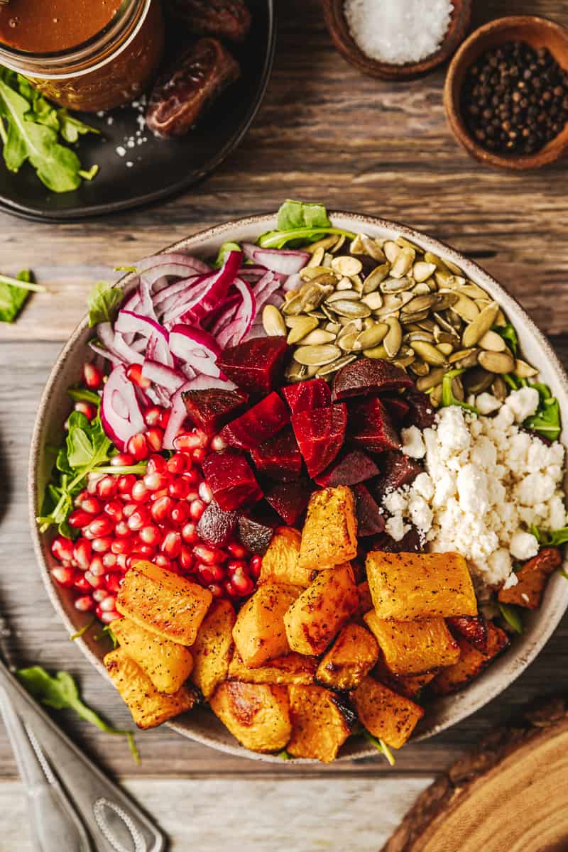 An overview shot of a bowl of a roasted butternut and beetroot salad on a wood background near utinsels and dressing