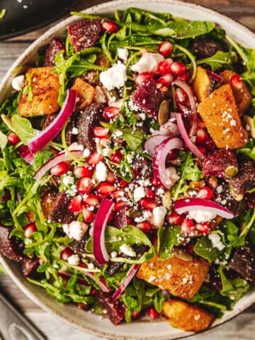 An overview shot of a bowl of a roasted butternut and beetroot salad on a wood background near utinsels and dressing