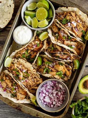 An overview shot of instant pot carnitas in tacos next to onions, limes, and sour cream on a wood backrgound