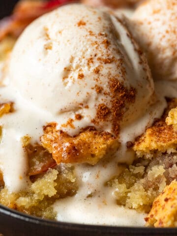 Peach cobbler in a cast iron skillet topped with a scoop of melting ice cream and cinnamon.