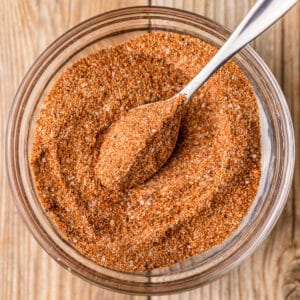 A small glass bowl filled with a sweet and smokey chicken dry rub along with a small silver spoon on a wood background.