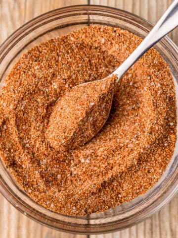 A small glass bowl filled with a sweet and smokey chicken dry rub along with a small silver spoon on a wood background.