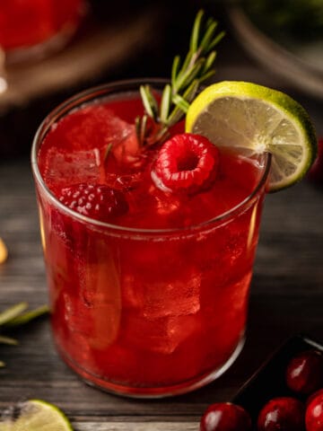 A glass filled with a cranberry raspberry mocktail topped with raspberries, rosemary, and limes on a wood background