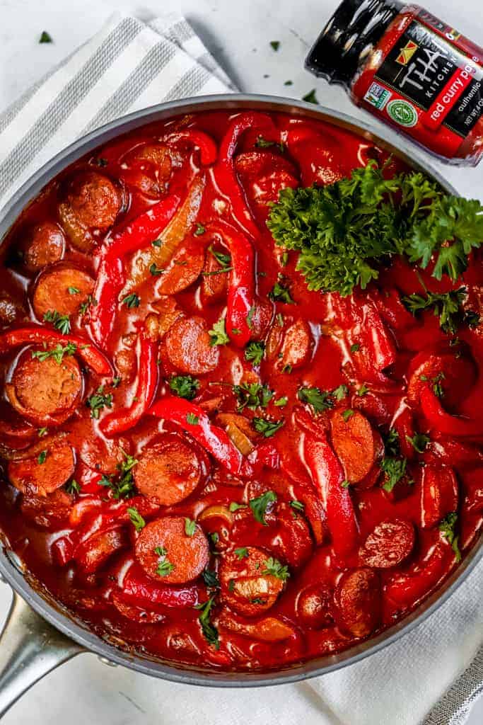 An overview of a pan of coconut curry with sausage and peppers next to a jar of thai kitchen red curry paste