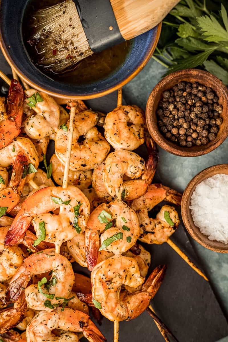 An overview shot of smoked shrimp skewers on a black serving plate next to shrimp marinade and garnishes