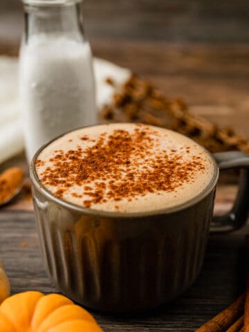 A grey mug filled with a paleo pumpkin spice latte on a wood background next to cinnamon sticks and pumpkins.