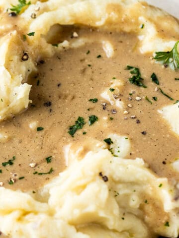 A bowl of mashed sweet potatoes with a pool of gravy and topped with parsley, salt and pepper on a wood background.