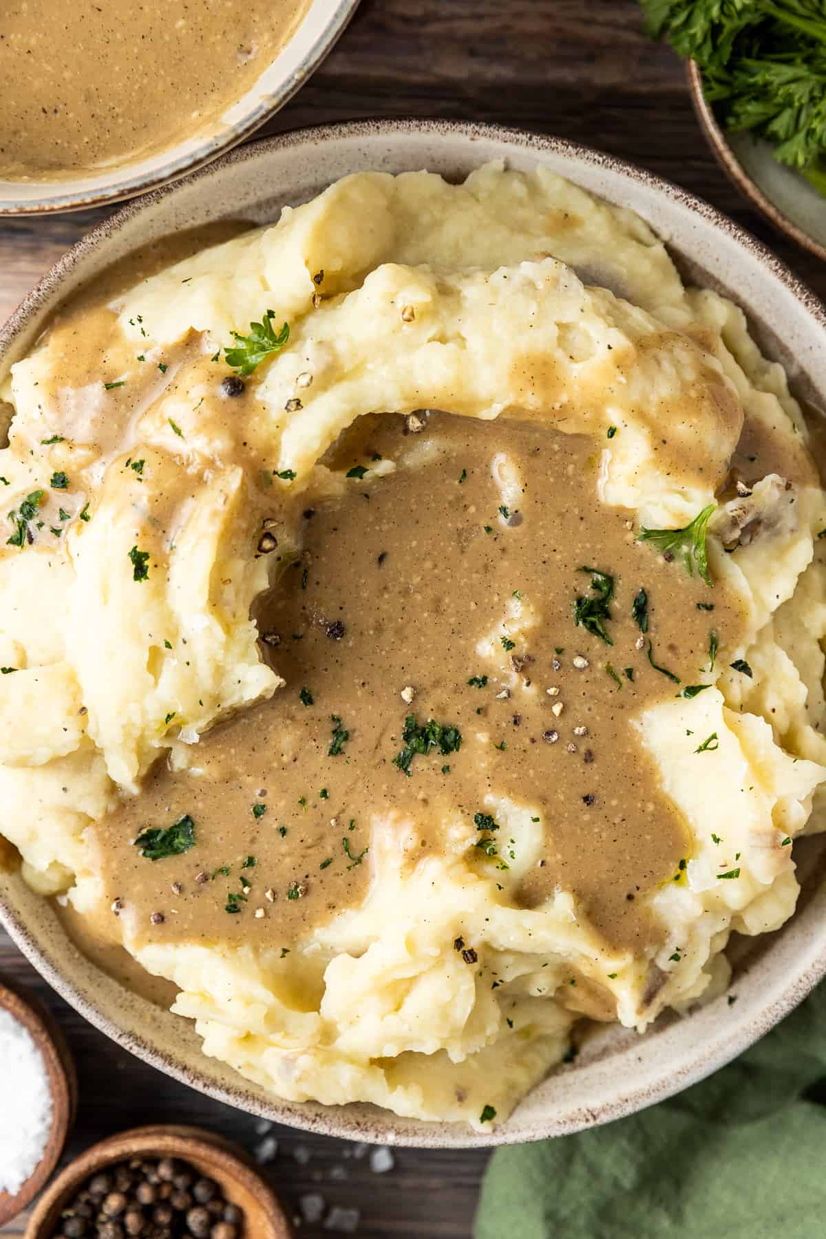 A bowl of mashed sweet potatoes with a pool of gravy and topped with parsley, salt and pepper on a wood background.