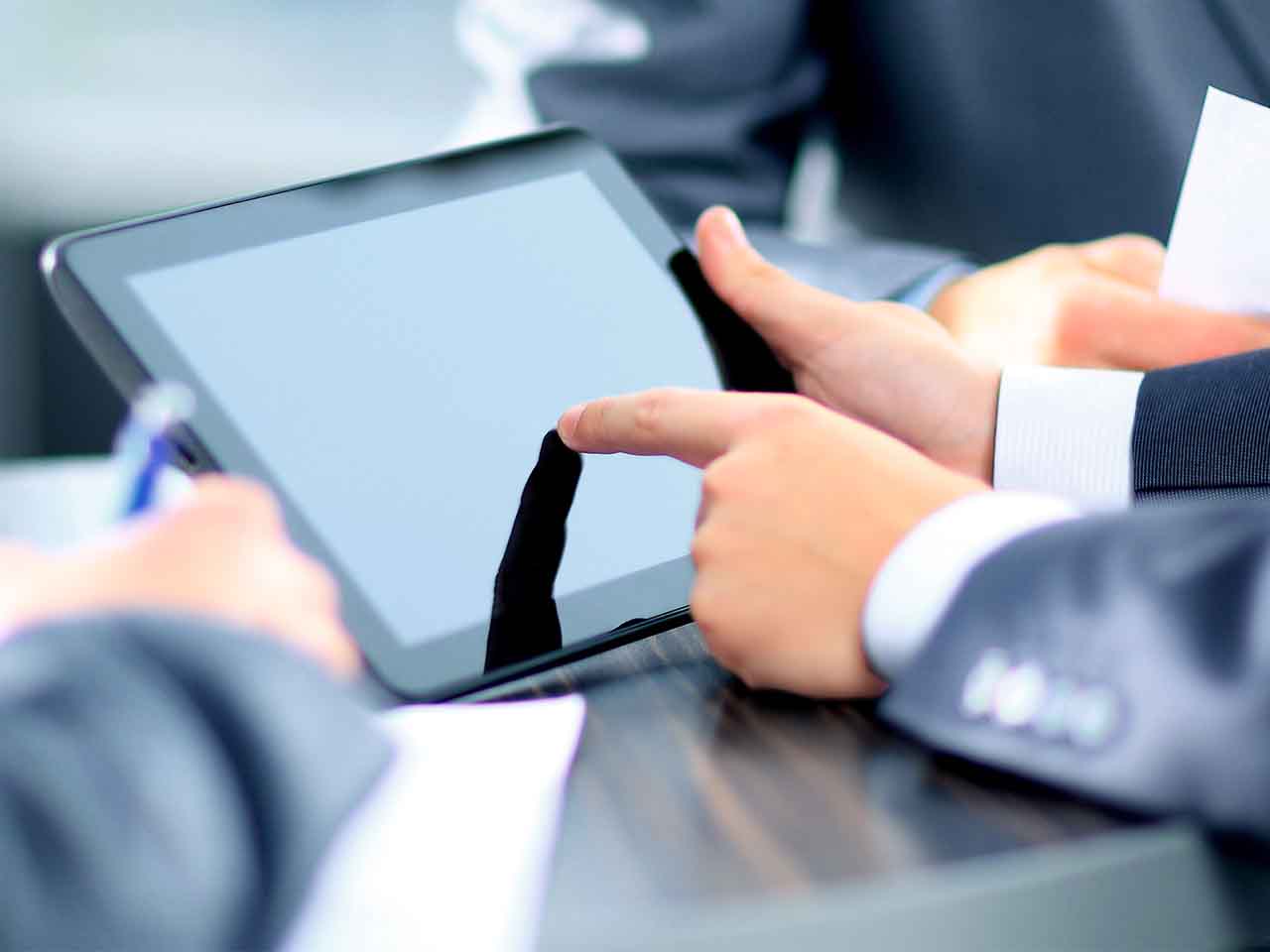 Close-up of business professionals using a digital tablet during a meeting, with one person pointing at the screen while another takes notes.