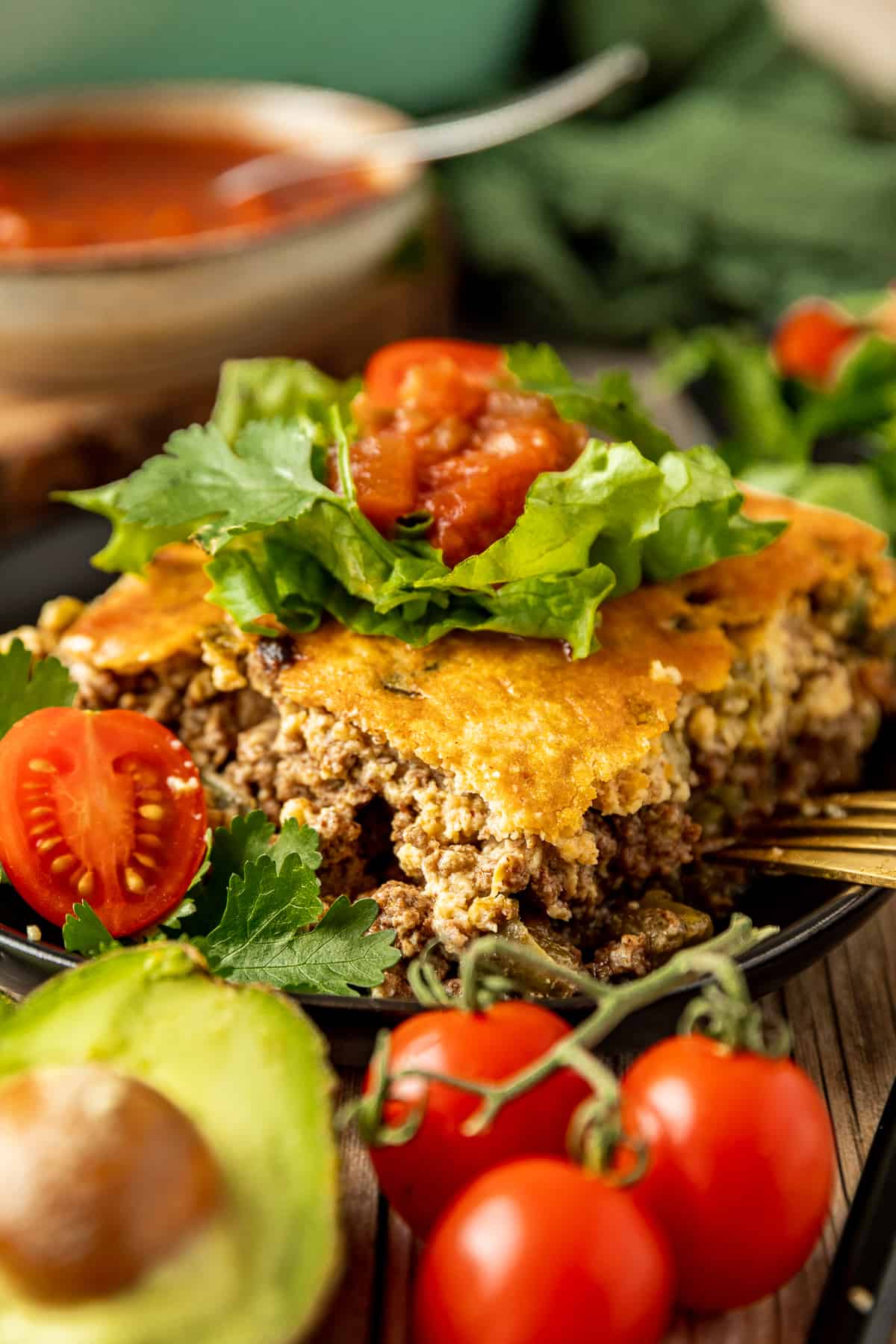 A square piece of taco casserole topped with tomatoes, avocado, and lettuce on a black plate.