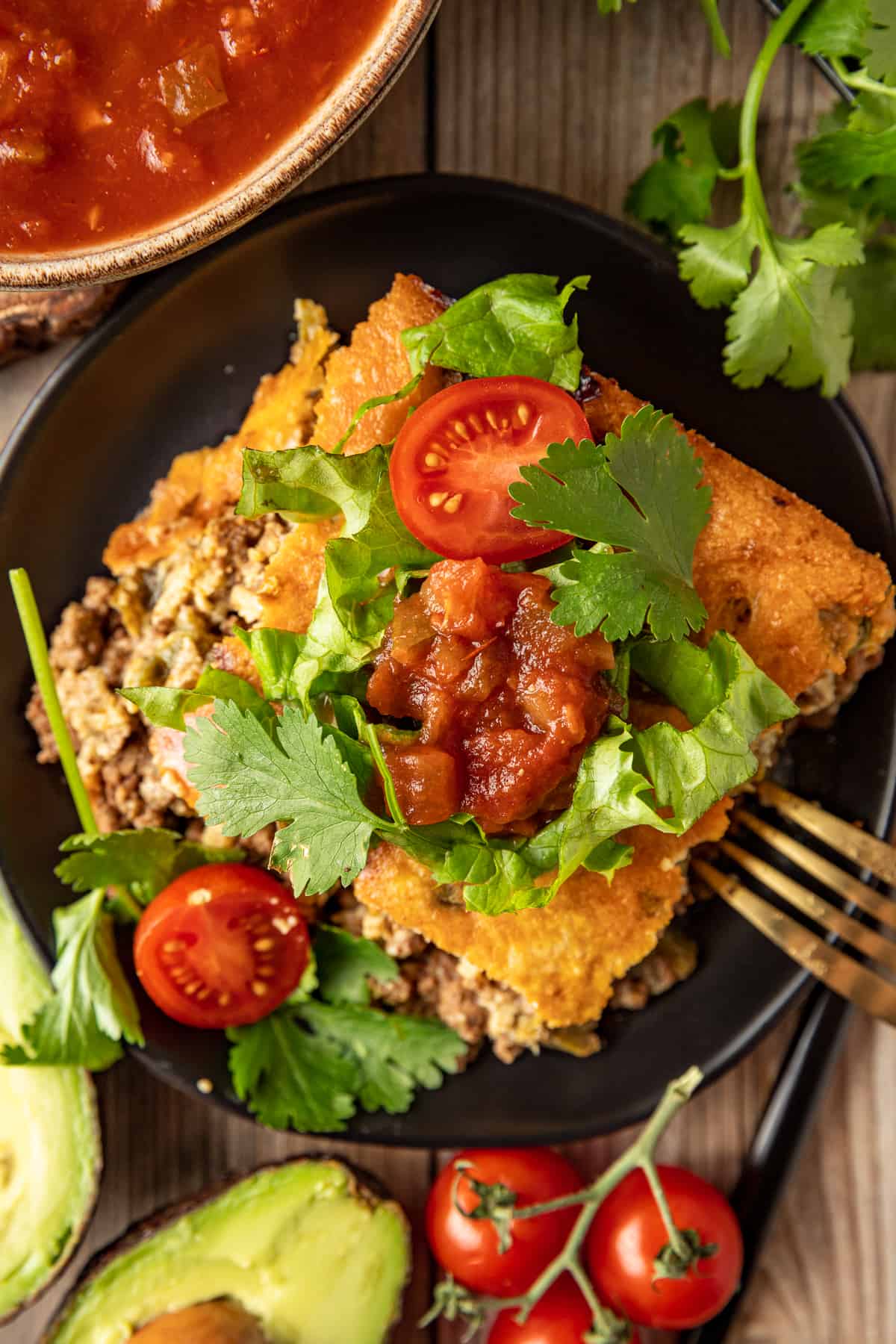 An overview shot of a square piece of taco casserole topped with tomatoes, avocado, and lettuce on a black plate.