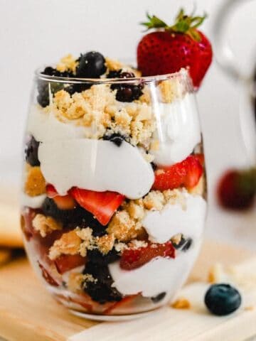 A glass with layers of berries, whipped coconut cream, and shortbread cookie crumbles on a cutting board next to cookies and berries.