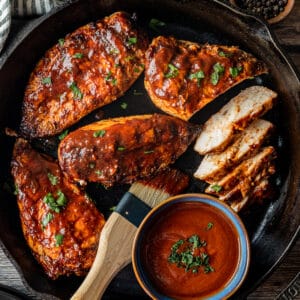 An overview shot of juicy BBQ chicken in a cast iron pan next to a bowl of BBQ sauce topped with chopped parsley.