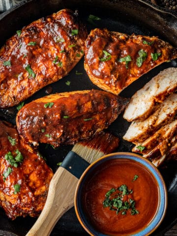 An overview shot of juicy BBQ chicken in a cast iron pan next to a bowl of BBQ sauce topped with chopped parsley.