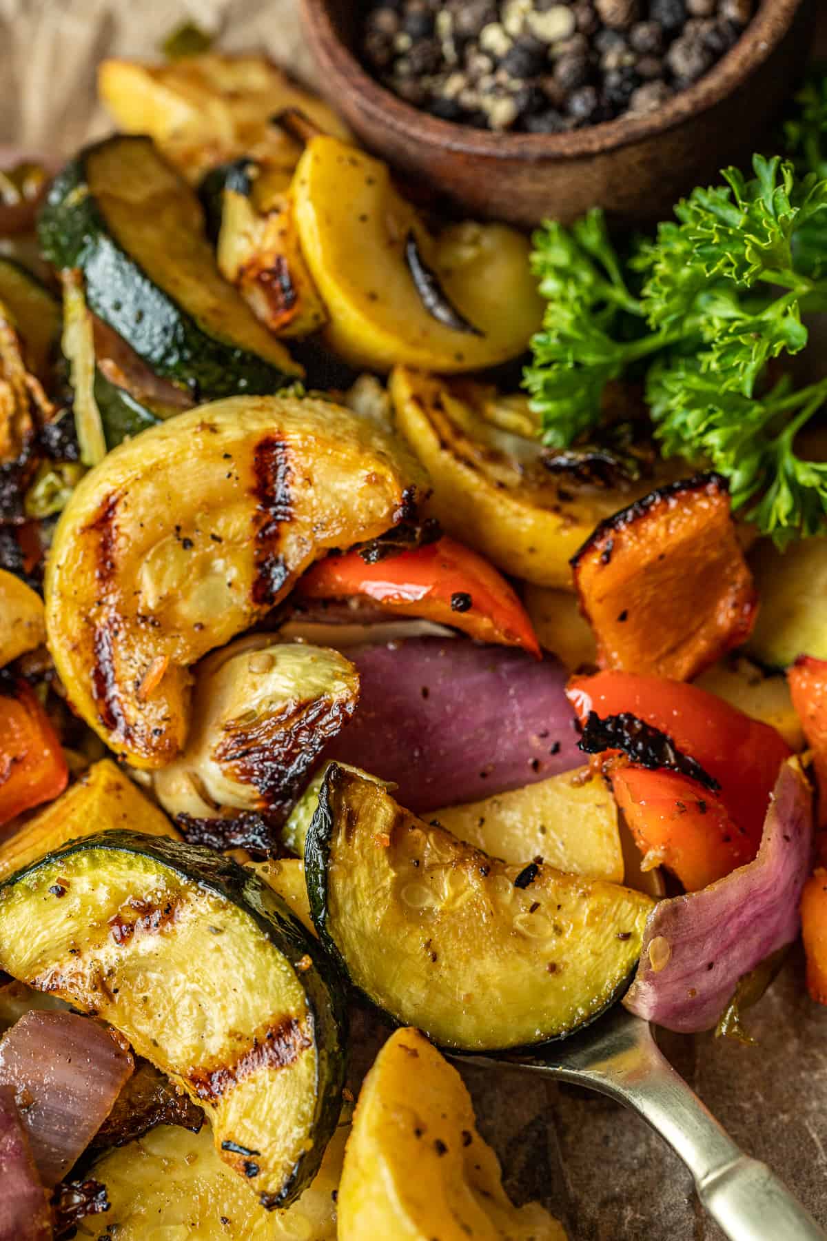 Smoked vegetables with grill marks being scooped by a gold spoon near parsley and peppercorns.