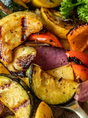 Smoked vegetables with grill marks being scooped by a gold spoon near parsley and peppercorns.