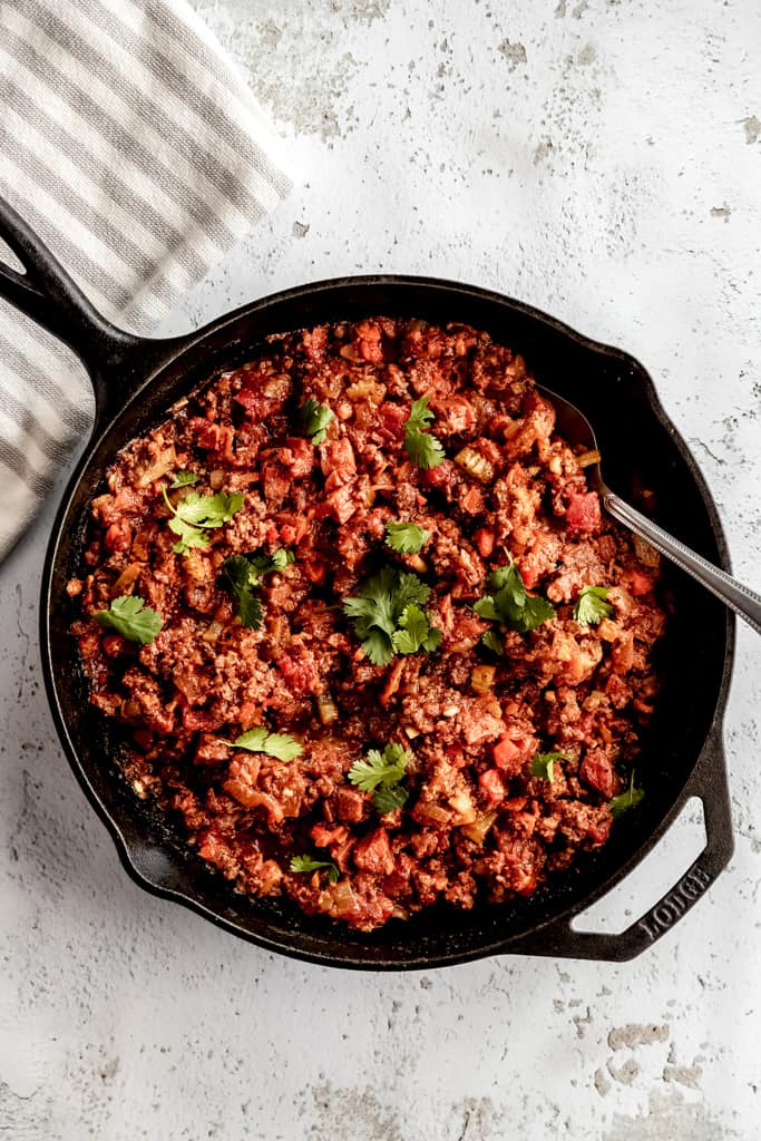 A cast iron pan of spaghetti meat sauce on a grey background next to a towel. 