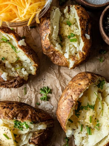 An overview shot of smoked baked potatoes filled with melted butter, chives, and parsley next to a bowl of shredded cheese.