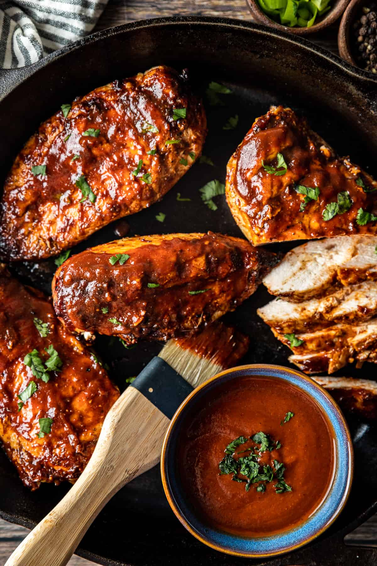 An overview shot of juicy BBQ chicken in a cast iron pan next to a bowl of BBQ sauce topped with chopped parsley.