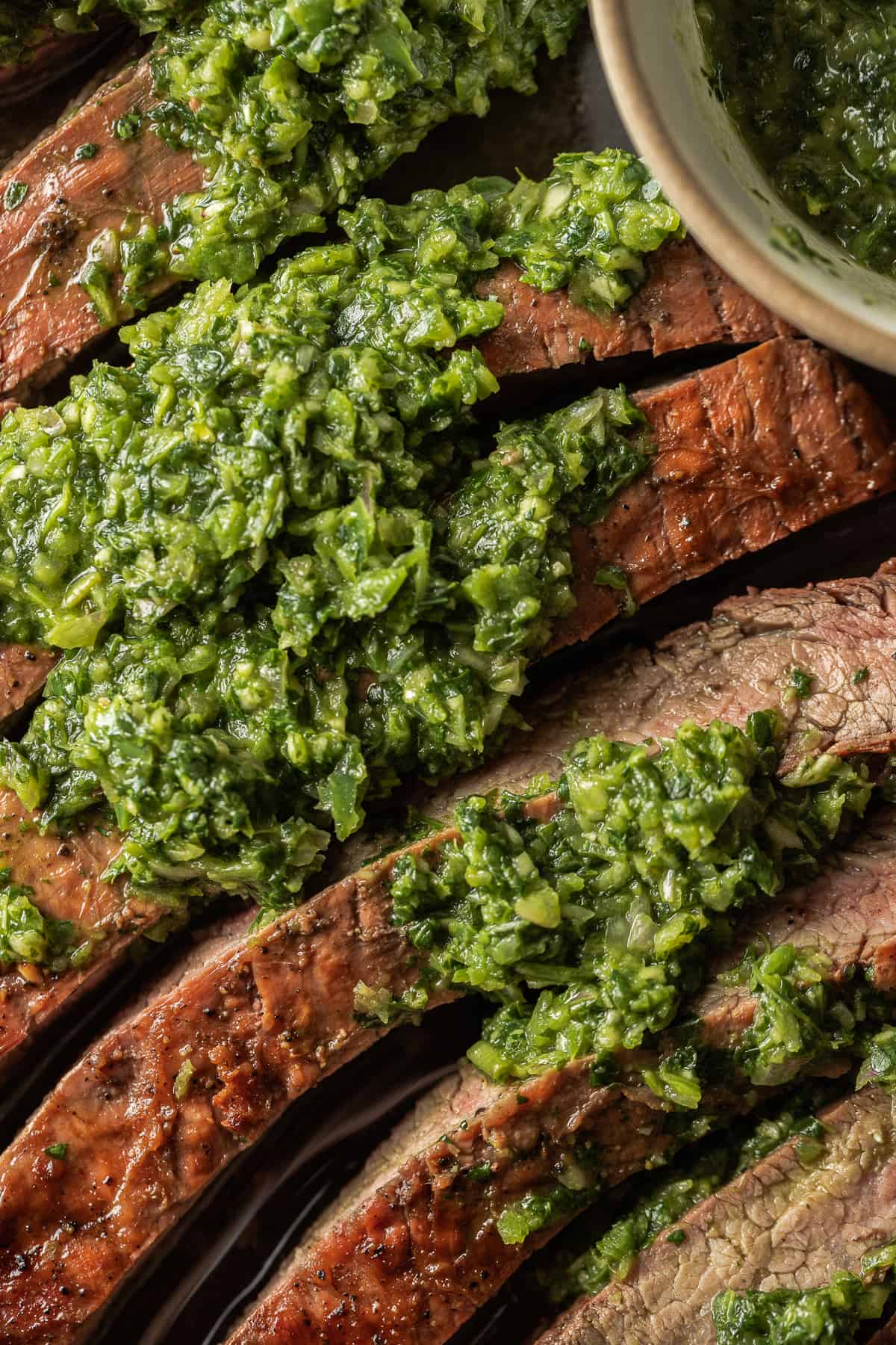 A sliced grilled flank steak topped with chimichurri next to a bowl of chimichurri.