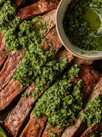 A sliced grilled flank steak topped with chimichurri next to a bowl of chimichurri.