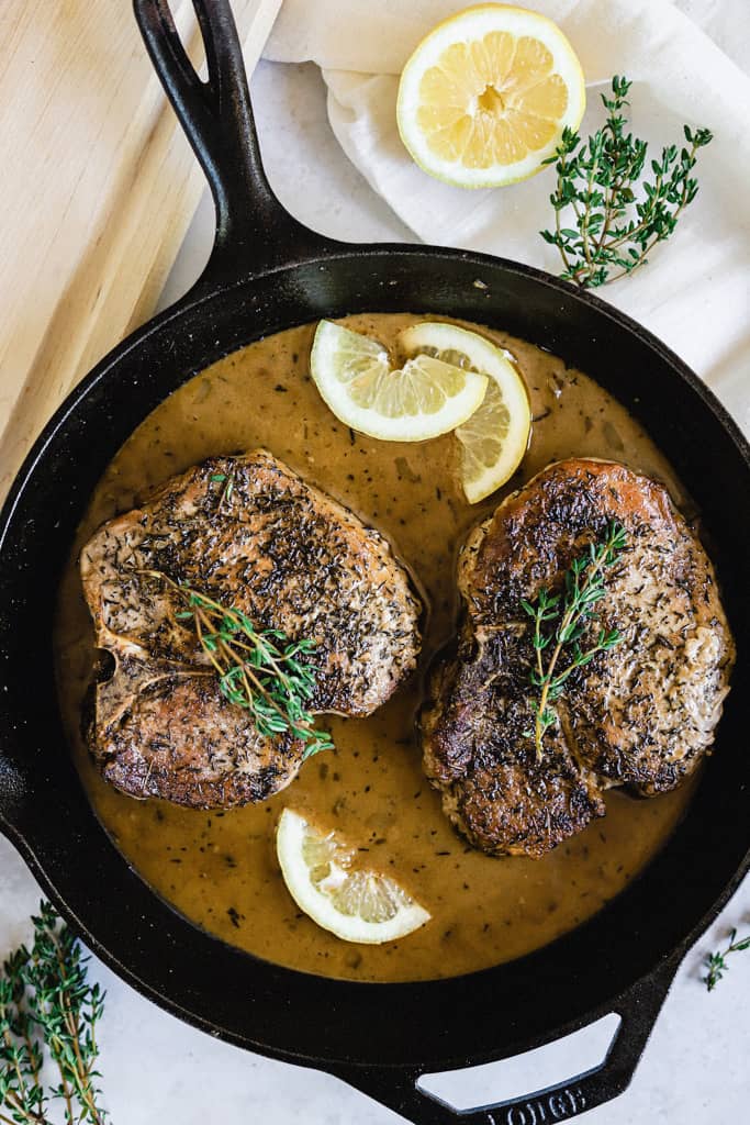 Two Pork Chops in a skilled topped with thyme and lemon wedges next to a cutting board and linen