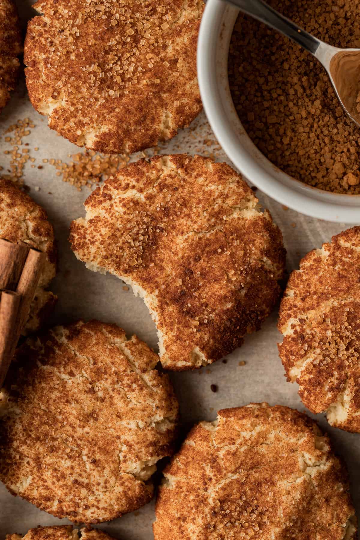 An overview shot of snickerdoodle cookies, with a bite removed, on a brown surface near a bowl of cinnamon sugar. 