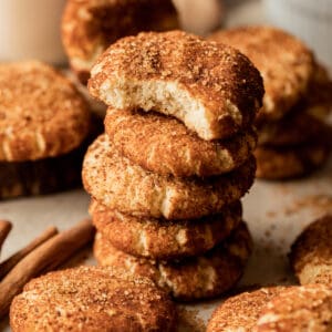 A stack of vegan snickerdoodle cookies with a bite taken out on a brown background near cinnamon sticks