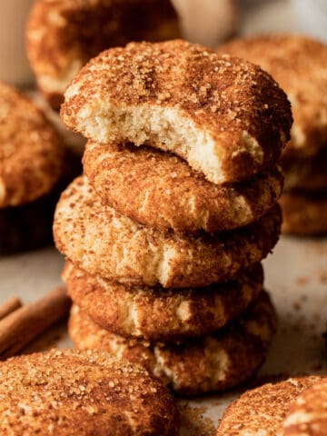 A stack of vegan snickerdoodle cookies with a bite taken out on a brown background near cinnamon sticks