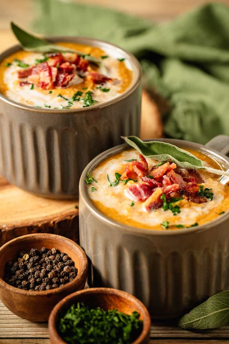 Two bowls of Whole30 Butternut Squash Soup topped with coconut cream, bacon, and fresh sage on a wood background