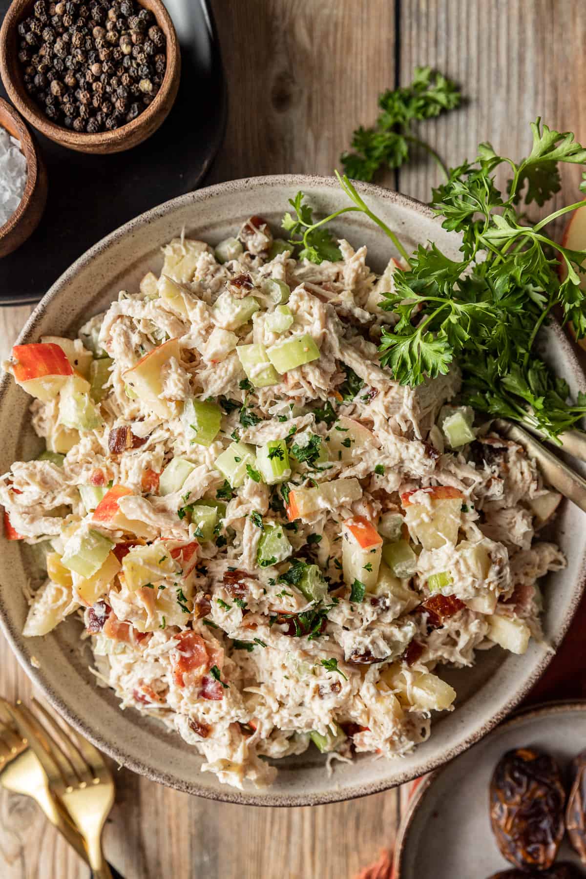 Chicken salad in a bowl with on a wood background near dishes of salt, pepper, and dates.